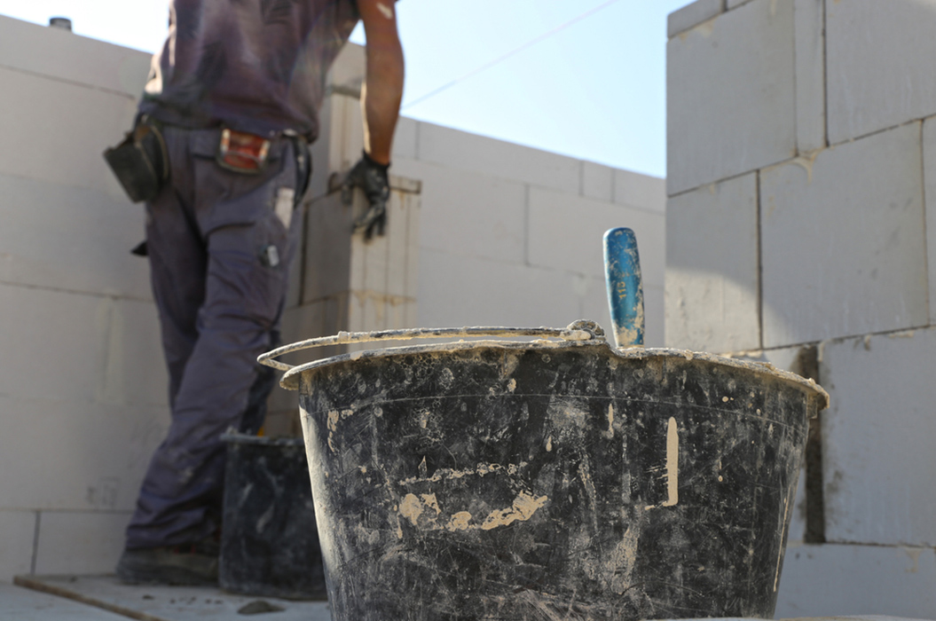 Arbeiter auf Baustelle, konzentriert, schwarze Kelle im Vordergrund, Mauer im Hintergrund, sonnig.