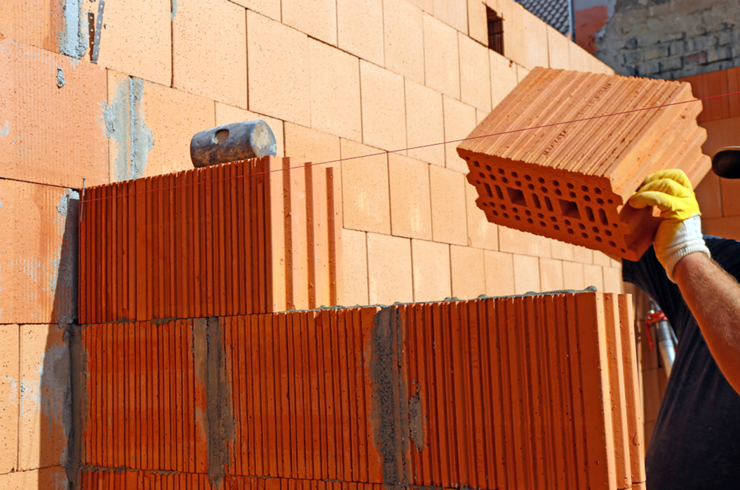 Person legt mit gelben Handschuhen rote Ziegelsteine auf eine Mauer; Ziegelstruktur gut sichtbar.