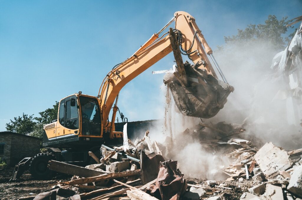 Destruction of old house by excavator. Bucket of excavator breaks concrete structure.